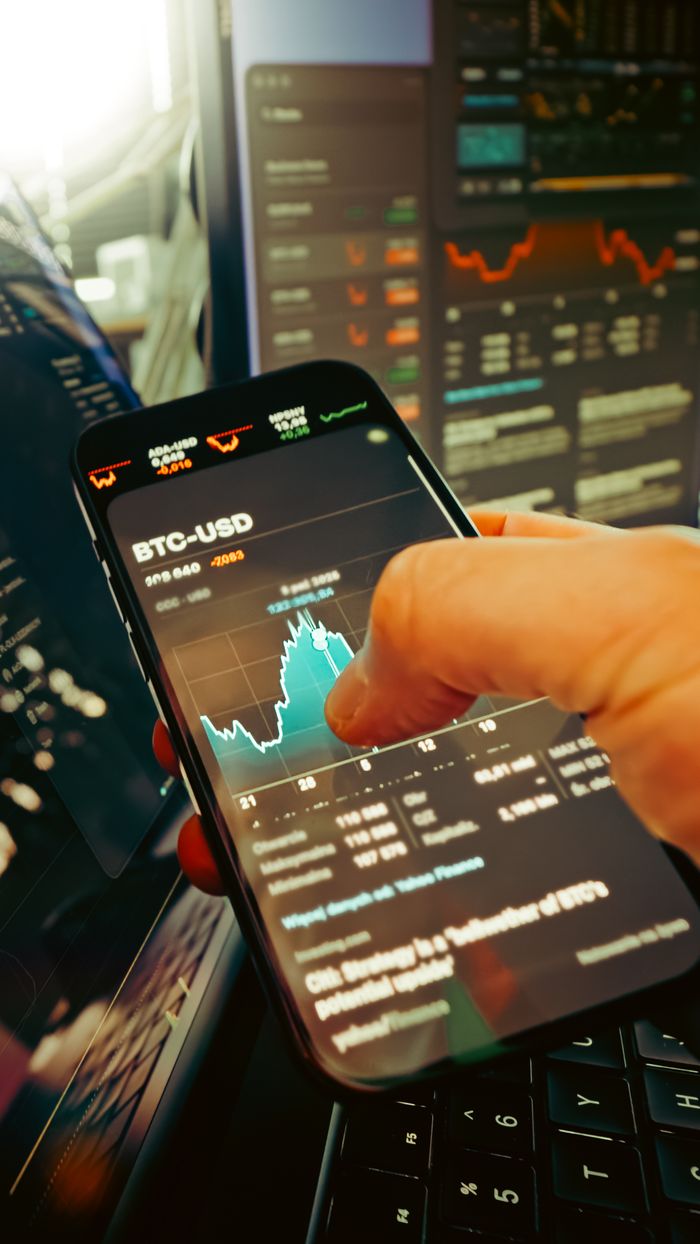 hero-img-01 A hand holds a smartphone displaying BTC-USD chart amidst digital trading screens, highlighting crypto finance.